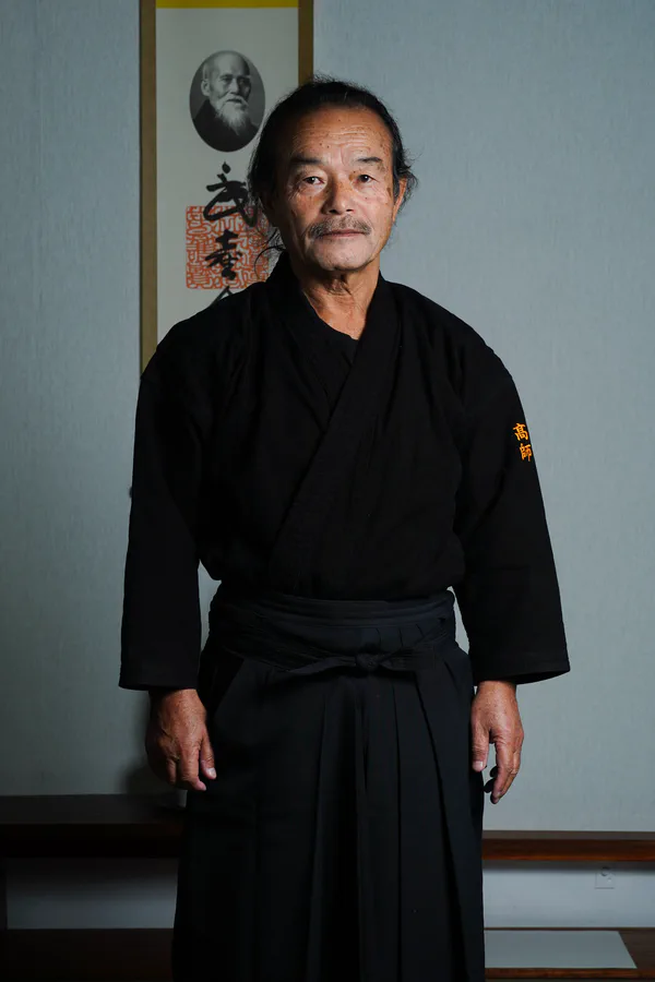 Saburo Takayasu Sensei in formal dress, before a scroll bearing O-Sensei's portrait.